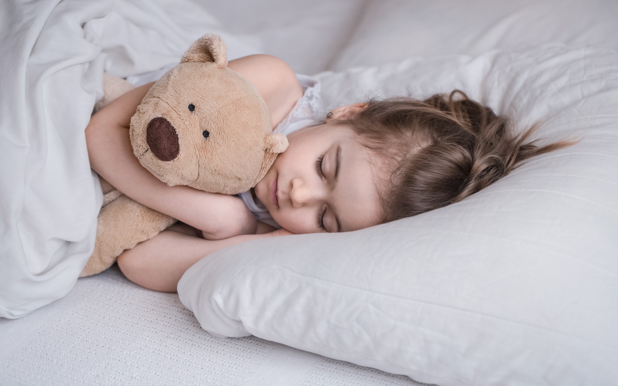 Lille pige ligger i seng med hvidt sengetøj og sover, mens hun krammer en lysebrun bamse