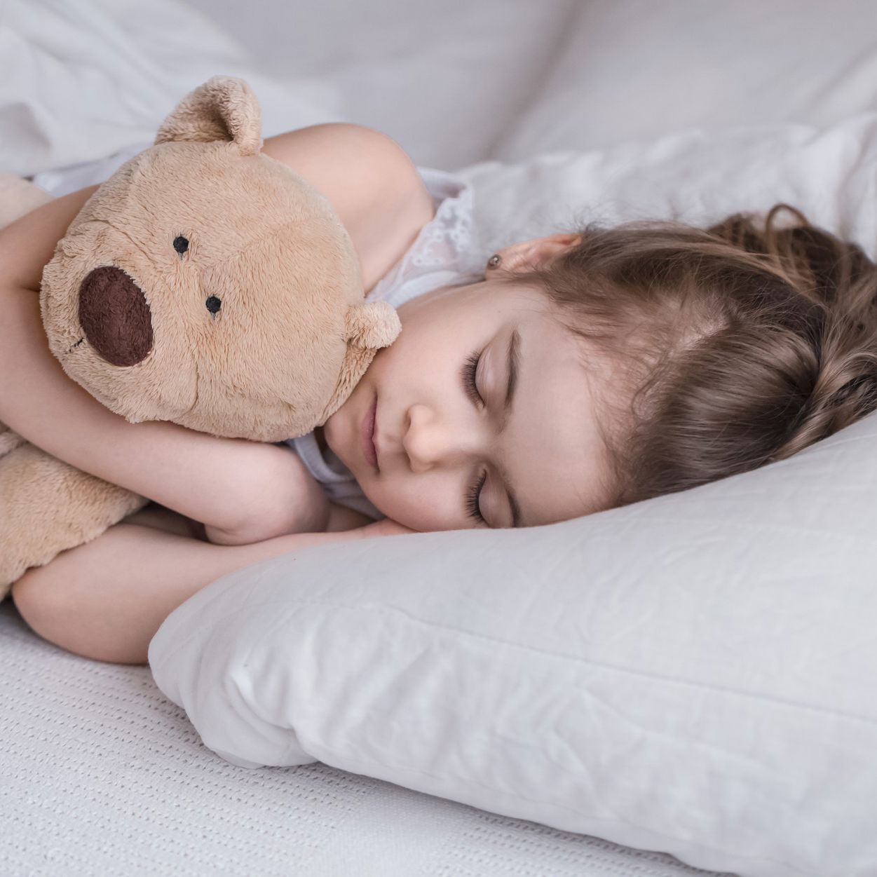 Lille pige ligger i seng med hvidt sengetøj og sover, mens hun krammer en lysebrun bamse