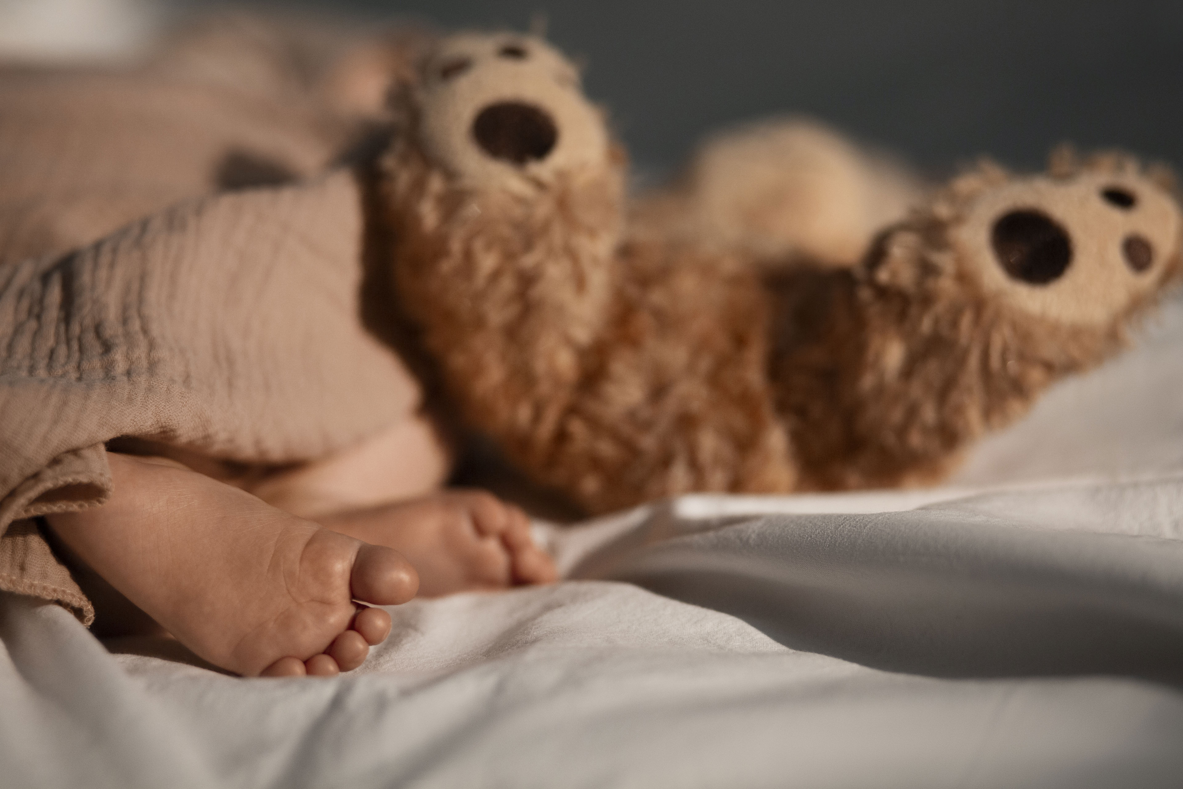 Baby fødder der stikker ud under tæppe. Brun bamses fødder ligger og stritter i vejret ved siden af baby.