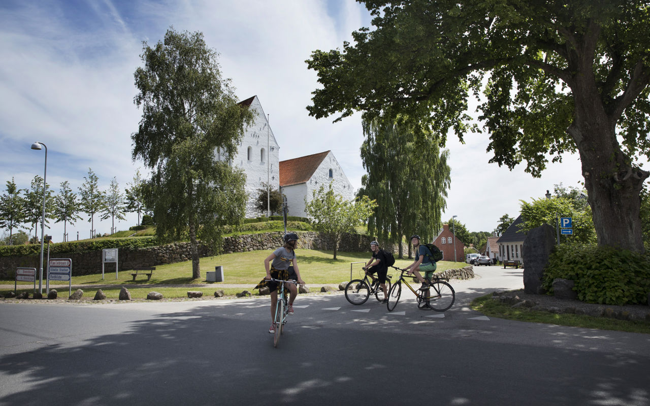 tre cykelister foran hvid kirke i grønne omgivelser